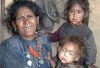 Woman in Humla holding two young children in worn clothing inside their home.