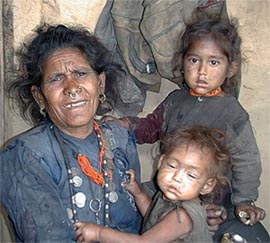 Woman in Humla holding two young children in worn clothing inside their home.